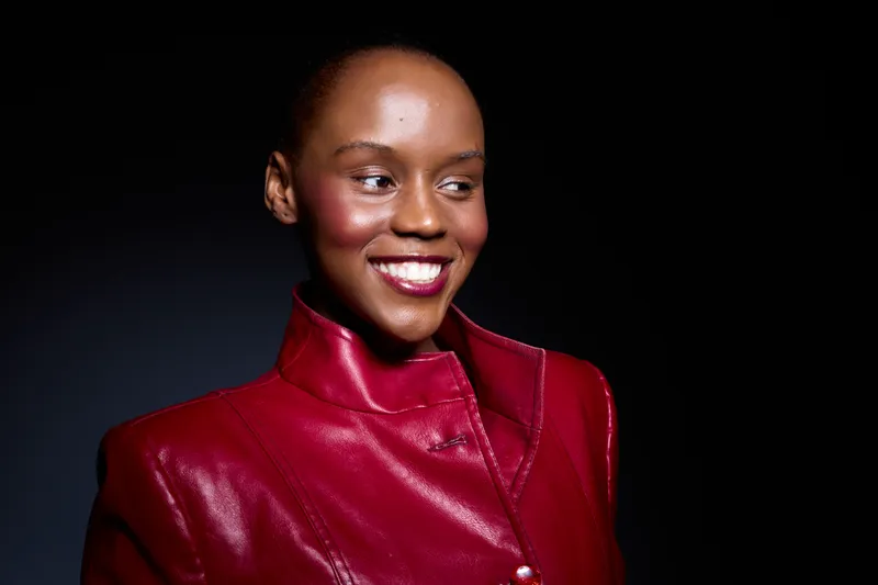 Smiling young black woman with dark skin wearing shiny red leather jacket looks sideways. Studio portrait on dark background. Concept of fashion, beauty, confidence, and style.