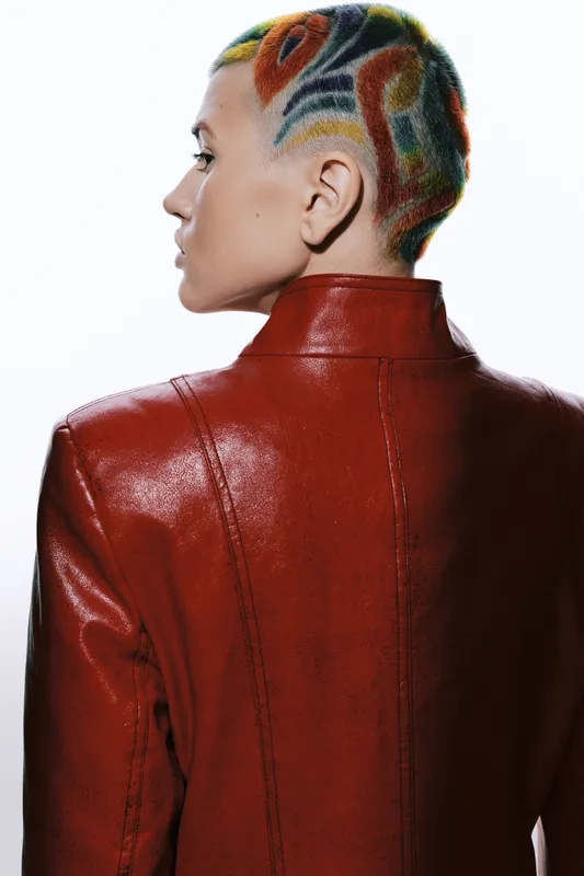 Woman with colorful shaved hair and intricate patterns wearing a red leather jacket, standing turned away with white background, showcasing bold style and creativity.