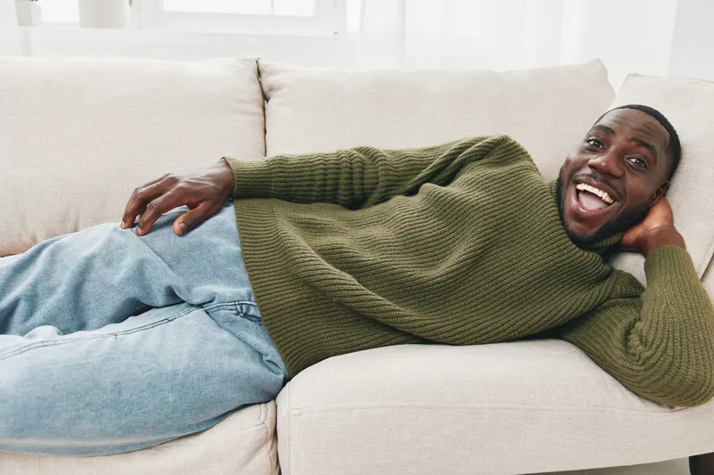 Cheerful young African man relaxing on a couch in a cozy sweater, embodying comfort and happiness in a modern home setting