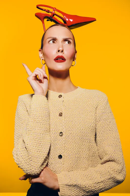 Young woman with red high heel shoe on head looks up with thoughtful expression. Bright yellow background and beige textured sweater create vibrant fashion concept.