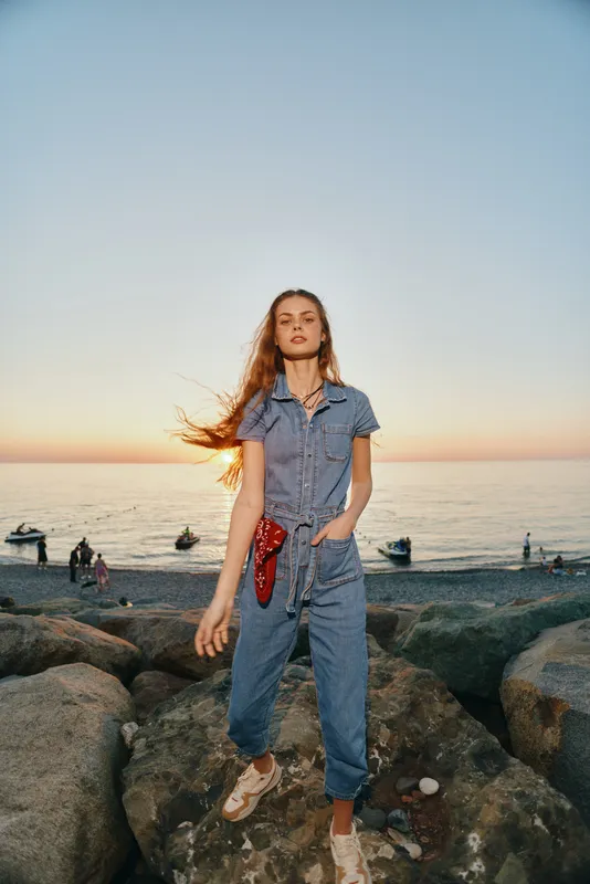 lifestyle woman in modern boho-western denim street style stands on rocks by the beach at sunset with a relaxed and confident mood in warm film color tones