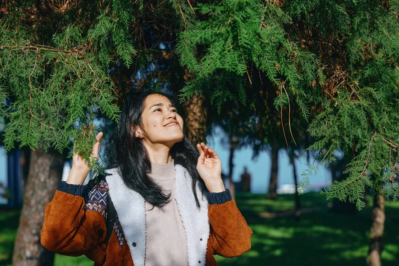 Woman under lush trees enjoying sunlight, smiling and looking up, outdoor moment of joy and relaxation in a park, casual jacket, fresh air, nature mood