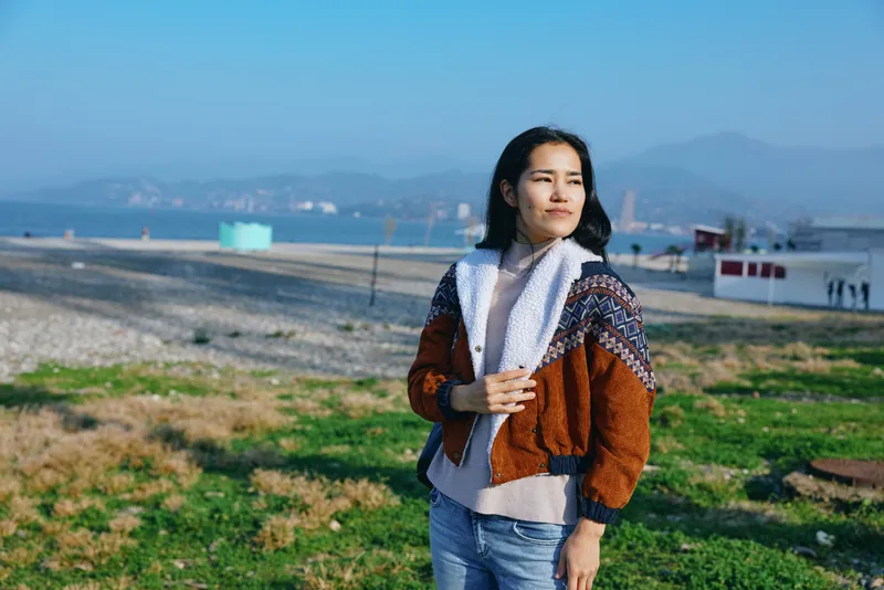 Woman wearing a warm scarf stands in a grassy field with distant mountains, casual outerwear, and a calm, thoughtful mood in an open outdoor landscape