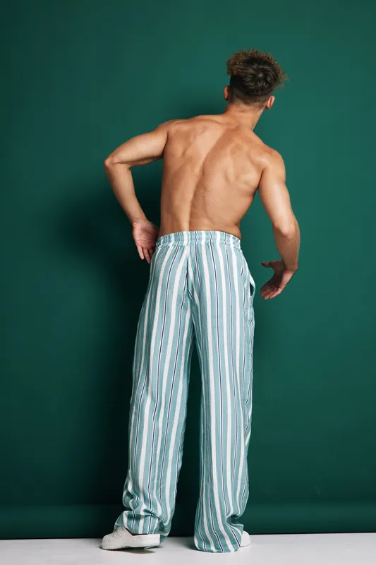Man with bare back wearing striped loose pants and white sneakers standing against green background in studio. Casual fashion, posture and style with modern clothing for young adult male.
