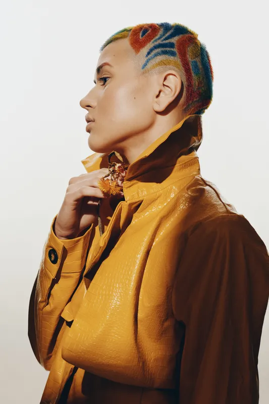 Woman with colorful patterned hair wearing a yellow leather jacket, posing in profile, showing stylish creative haircut and fashion-forward attitude in a studio setting.