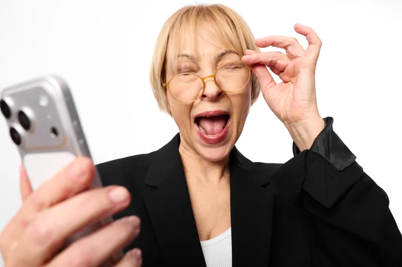 Woman laughing holding phone and adjusting glasses, elderly business portrait on white background, joyful smile, blazer suit, indoor isolated view close