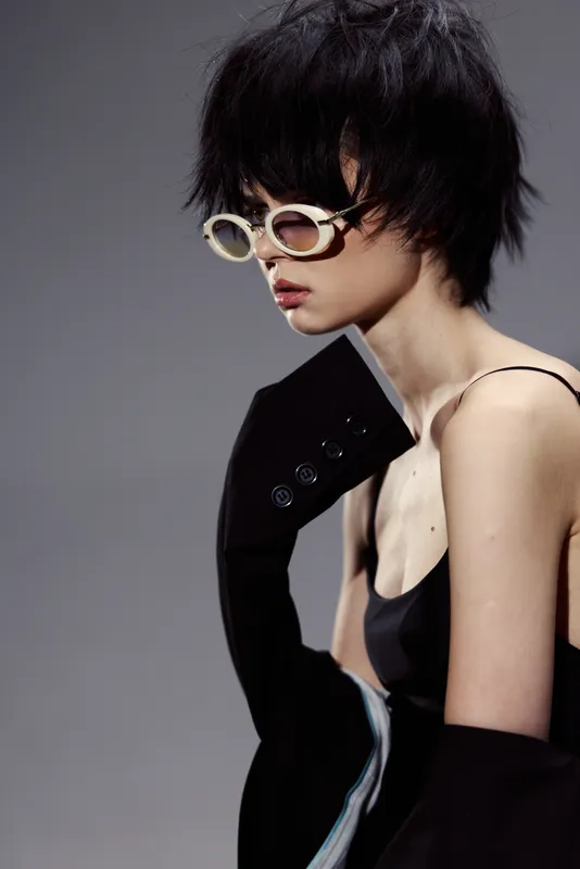 Stylish woman with short hair wearing sunglasses and modern black outfit in minimalist studio setting.