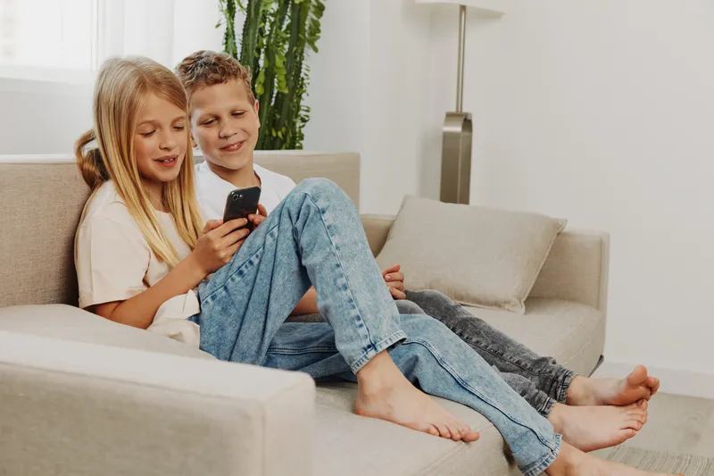 Two happy children sitting barefoot on a beige sofa sharing smartphone content in a bright living room with casual clothes and relaxed mood.