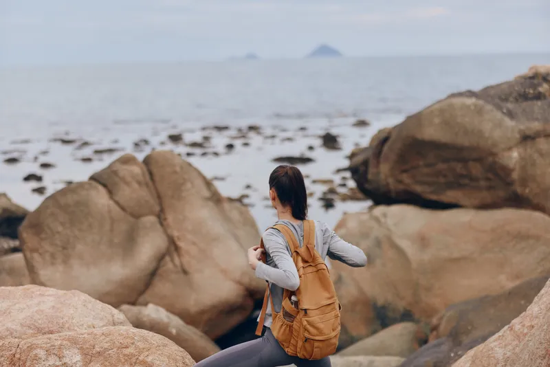 Young woman with brown backpack exploring rocky coast, showcasing adventure and serenity, wearing casual outdoor attire and enjoying nature