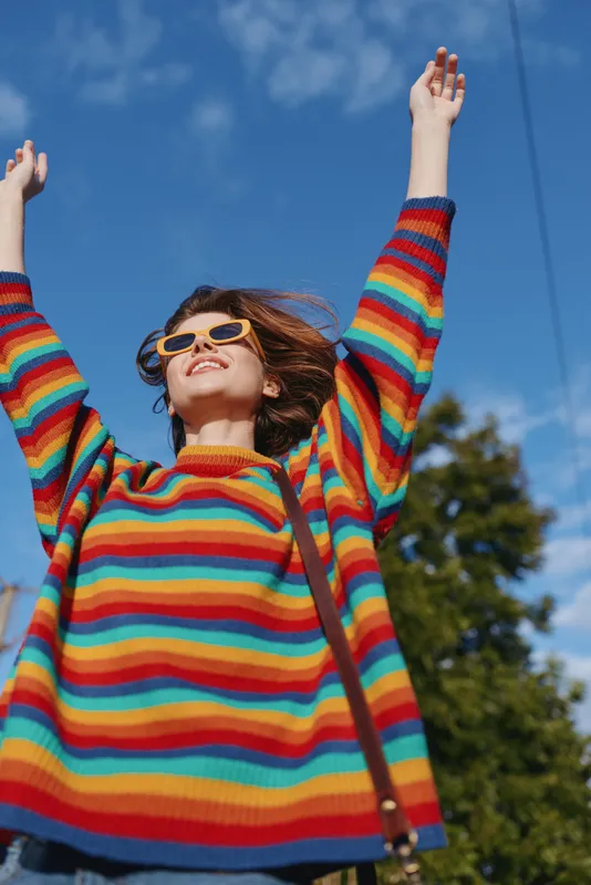 Woman in her 20s wearing a rainbow sweater and sunglasses, smiling with arms raised outdoors under blue sky, youthful lifestyle portrait, casual colorful fashion for travel and leisure.