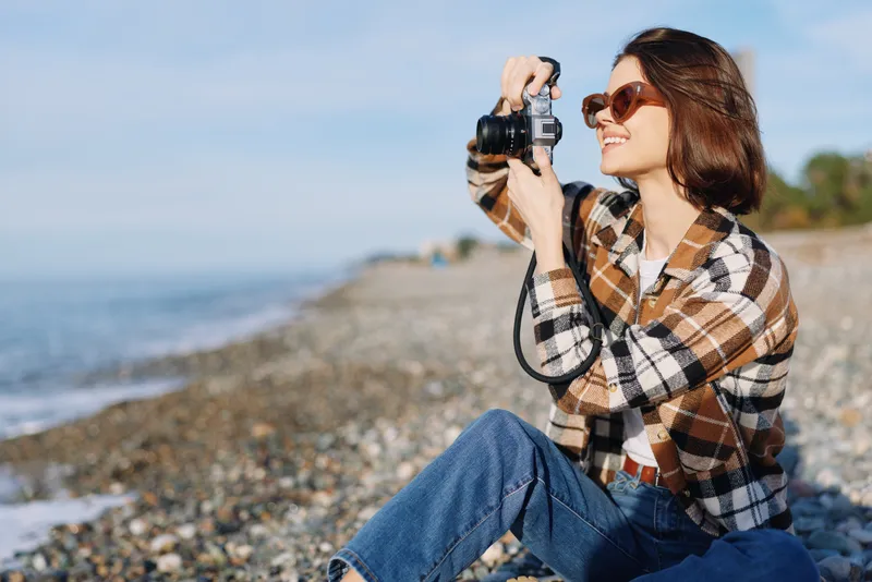 Casual woman at the beach using a camera for outdoor photography, sitting on pebbles with a plaid shirt, capturing seaside scenery and relaxed portrait moment.