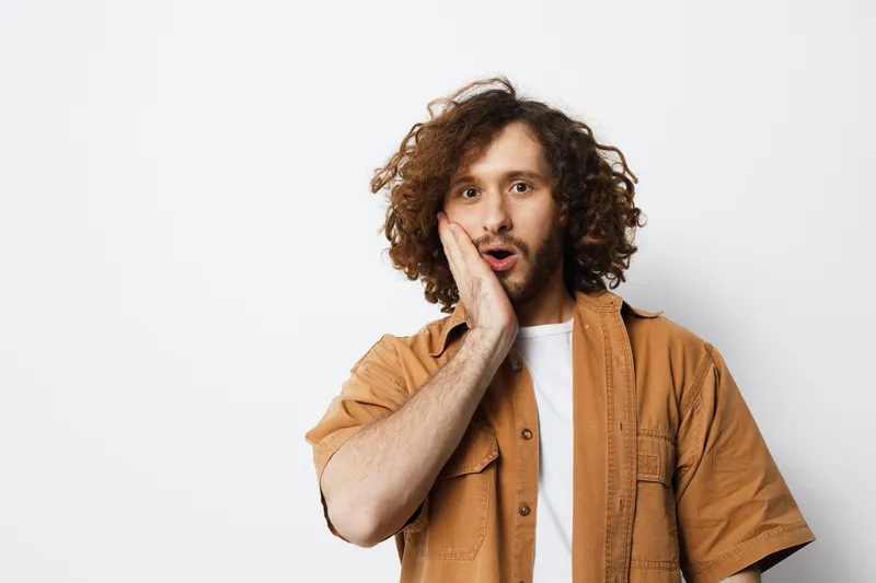 Surprised young man with curly hair in casual brown shirt expressing shock or amazement in a minimalist setting, ideal for emotional expression and lifestyle themes
