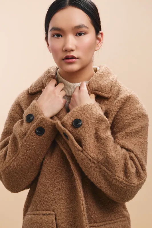 Asian woman wearing a cozy brown coat stands against a soft beige backdrop, projecting warmth and modern fashion. Her calm expression conveys confidence and approachable style in a studio portrait.