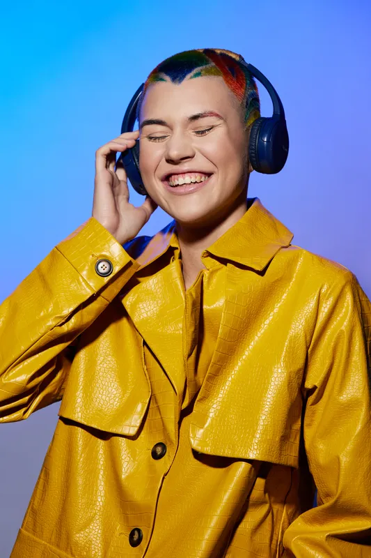 young woman with colorful hair enjoys music with headphones, wearing a yellow jacket against a vibrant blue background, expressing happiness and carefree mood.