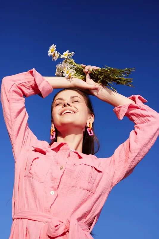 Joyful woman in a pink dress holding flowers, smiling against a clear blue sky, embodying themes of happiness and springtime freshness