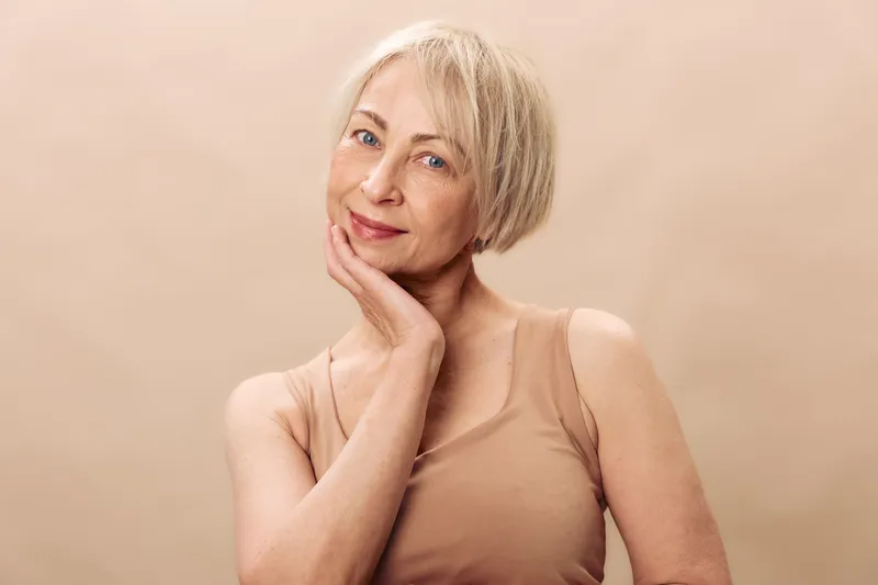 Portrait of a happy mature woman with short blonde hair touching her face gently. Soft beige background and natural makeup create a calm and confident mood. Concept of aging gracefully.