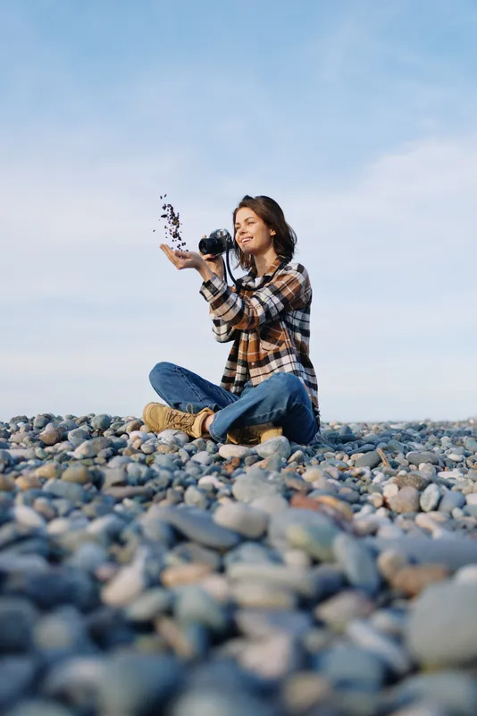 Woman on a pebble beach throws pebbles in the air, enjoying outdoor light, casual plaid shirt and denim, a moment of playful nature in a sunlit scene.