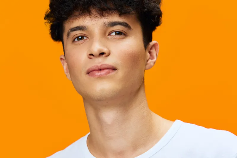 young man portrait with curly hair and smooth skin wearing white shirt, bright orange background, natural expression, casual style, studio lighting