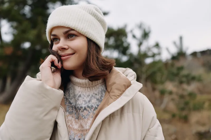 Woman talking on smartphone outdoors in winter, wearing a beige coat and knit hat, natural light, casual portrait in a rustic landscape