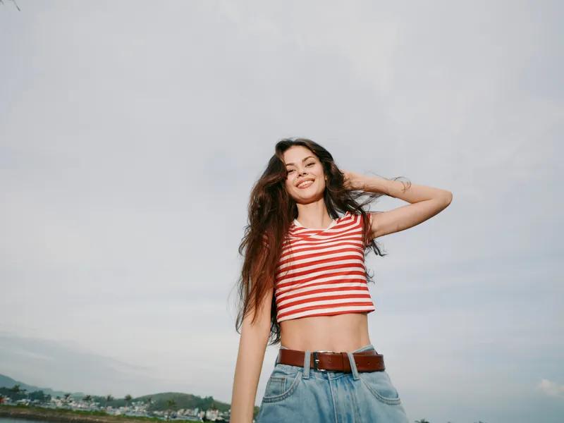 A young woman stands outdoors under a bright sky, wearing a red and white striped crop top and denim jeans. She smiles with carefree confidence.