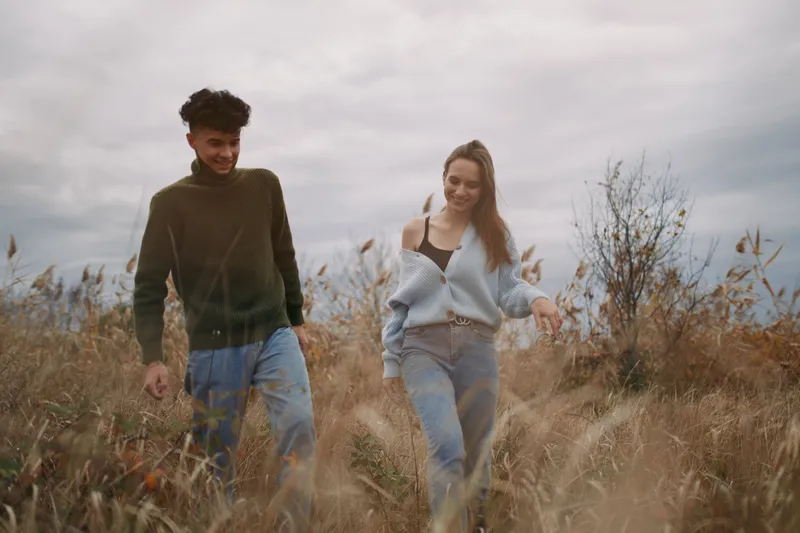 A young couple walking through tall grass in soft natural light, sharing genuine smiles and relaxed body language, capturing authenticity and trustworthiness in a candid, intimate moment.