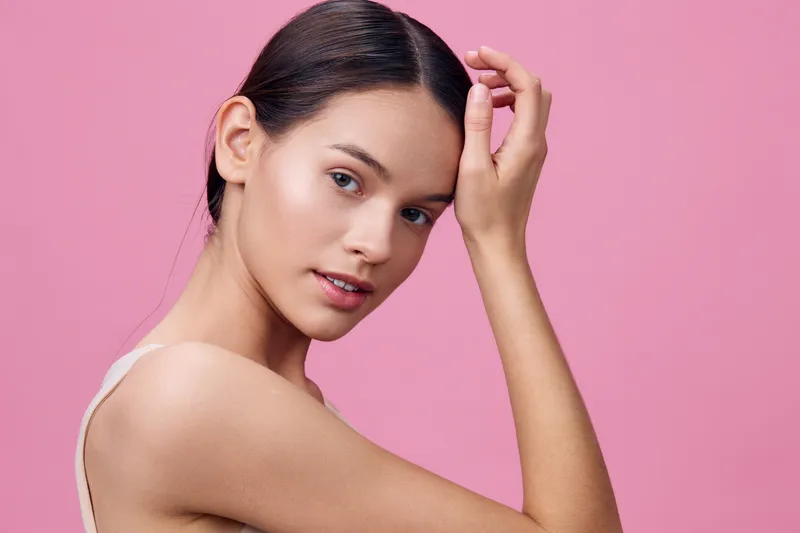 Beautiful young woman with a natural look and soft expression, posing against a pink background, perfect for beauty and skincare promotions