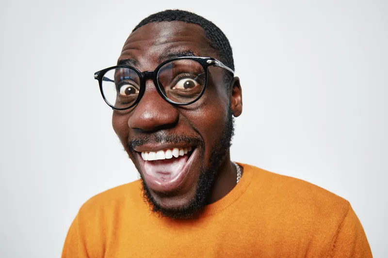African American man with dark skin wearing glasses and orange sweater smiling widely with excitement and joy on white background. Closeup portrait of happy black male.