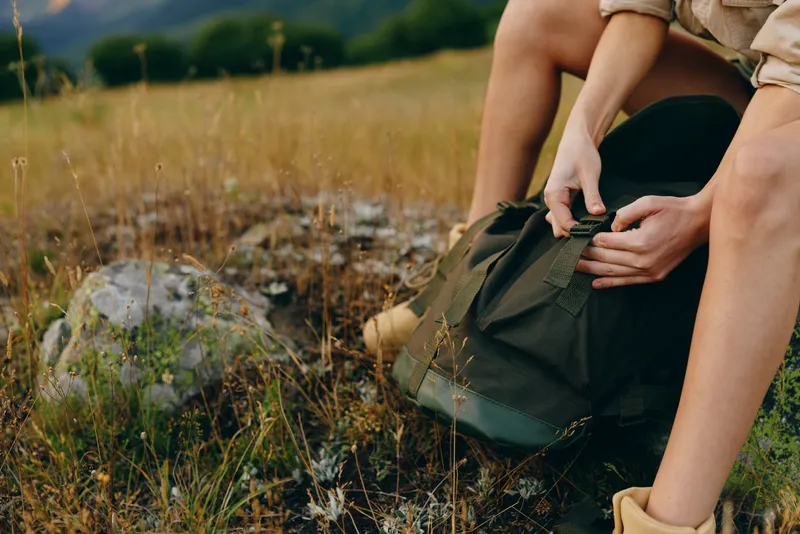 Young woman with rolled-up sleeves sits on a grassy field while opening a green backpack during a casual outdoor adventure.