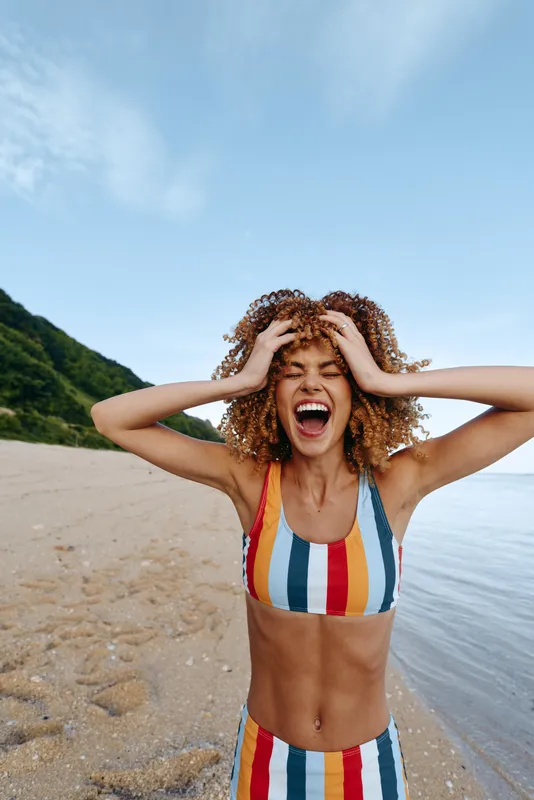 Laughing woman with curly hair on sandy beach by the sea, hands on head, striped swimsuit. Bright summer sunlight, outdoor vacation mood, blue sky and calm water, near green hills today.
