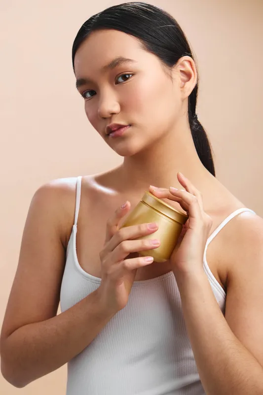 Asian woman stands in a neutral setting, holding a small jar with a calm smile, conveying focus, care, and a gentle beauty routine in soft light.