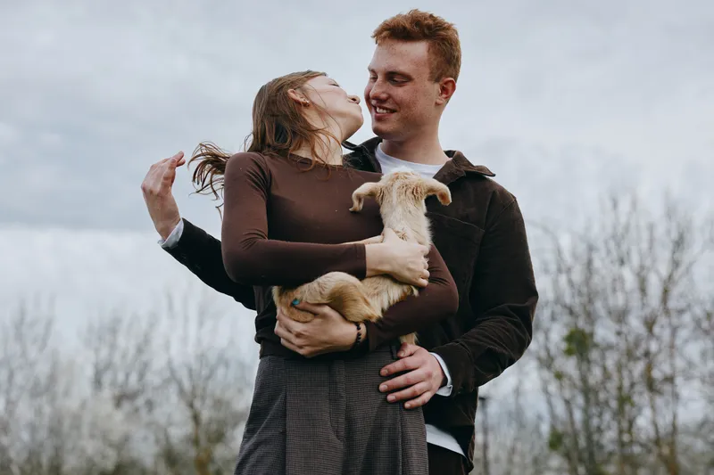 Young couple embraces outdoors while the woman holds a small goat in her arms, both smiling and sharing a tender moment in a natural lifestyle setting.