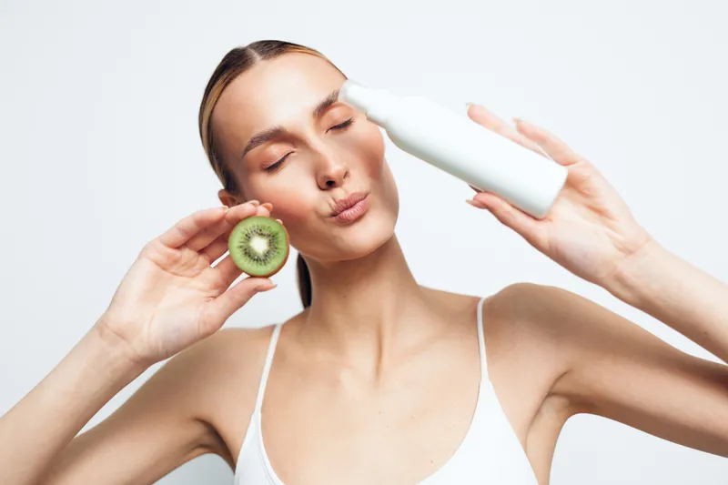 Young woman with clear skin holding a sliced kiwi and white cosmetic bottle, eyes closed, natural beauty concept, fresh skincare routine, bright studio background