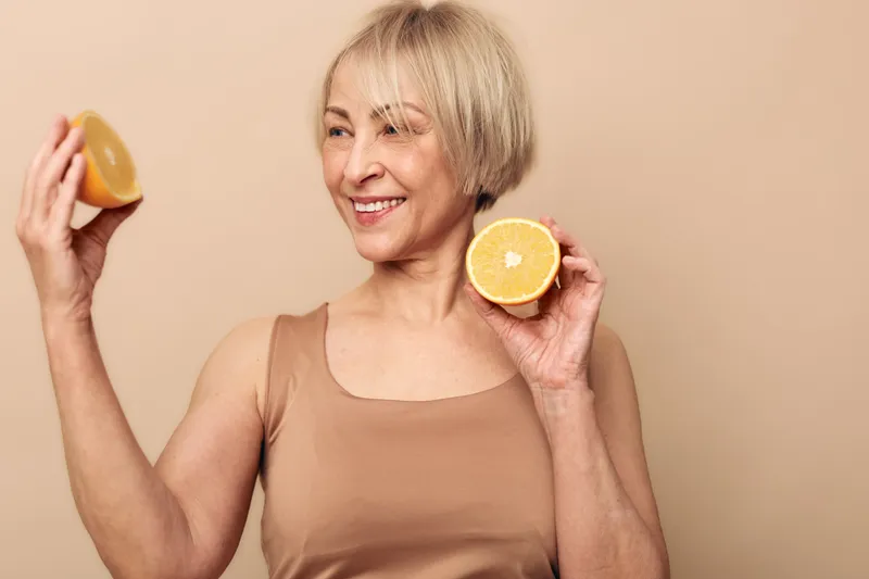 Smiling mature woman with short blonde hair holds two halves of fresh orange fruit against beige background. Concept of healthy lifestyle, vitamin intake, and natural beauty for seniors.