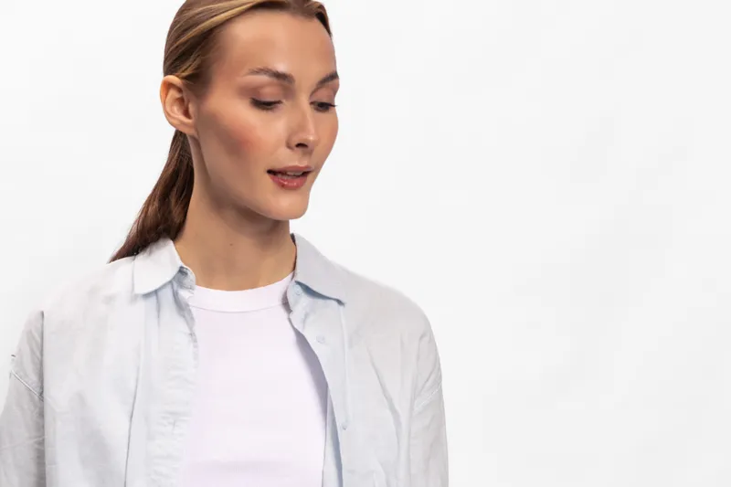 Young woman with light brown hair tied back wears white shirt and light jacket, looks downwards with calm expression. Studio portrait on white background with soft lighting.