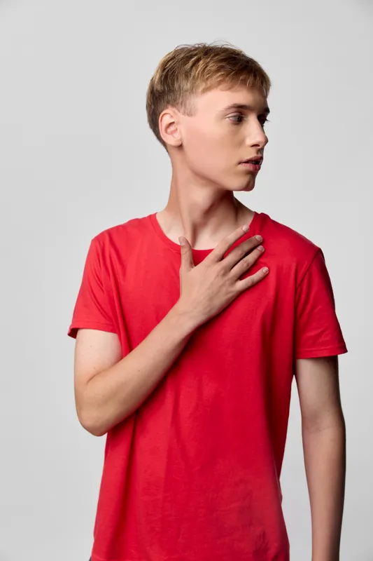 A young man wearing a red shirt places a hand on his chest with a thoughtful pose under studio lighting, conveying calm emotion and modern fashion sense in a clean minimal setting