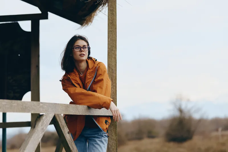 Woman lifestyle outdoors portrait orange jacket glasses wooden ladder sunlight, young woman leans on a wooden ladder by an outdoor structure, soft daylight, calm expression, shallow depth of field,