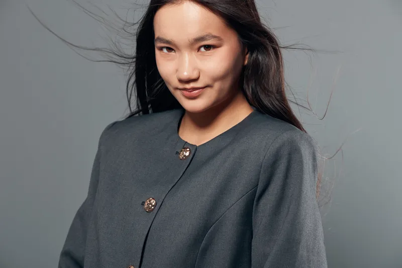 Young woman with long dark hair and slight smile wearing dark gray coat with buttons looking at camera on gray background. Studio portrait of confident female in fashionable outerwear.