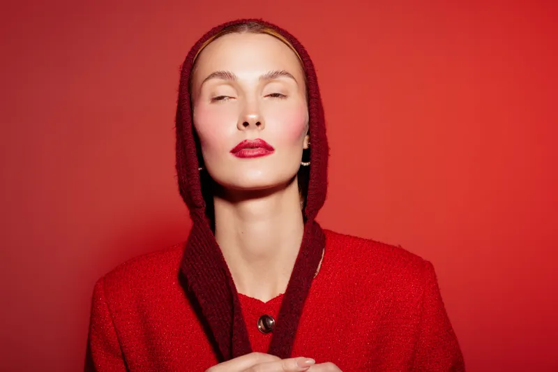 Portrait of a confident woman in red coat and hood with closed eyes against red background. Concept of fashion, elegance, beauty, and monochrome style in studio lighting.
