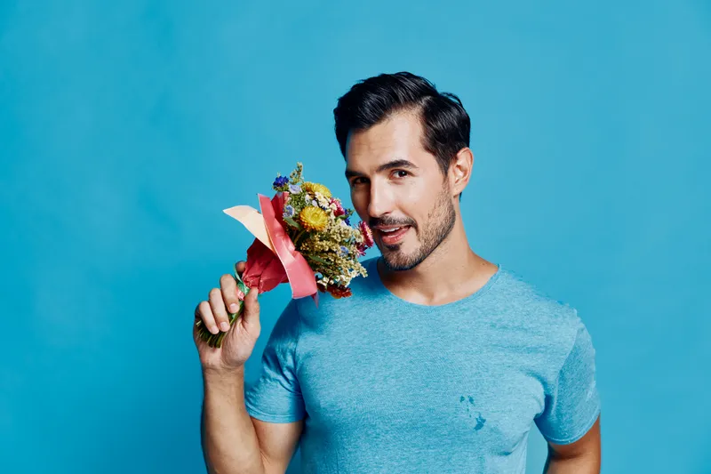 Handsome smiling man holding colorful bouquet of flowers, standing against a bright blue background. Celebrating love and special moments with joy.