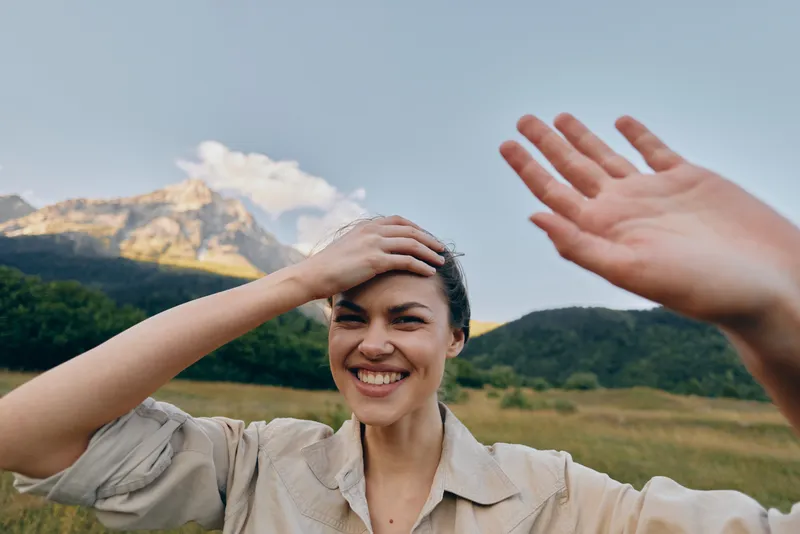 Young woman with short hair smiles broadly while shielding her eyes with one hand in a sunlit field with distant mountains, creating a bright, carefree moment outdoors