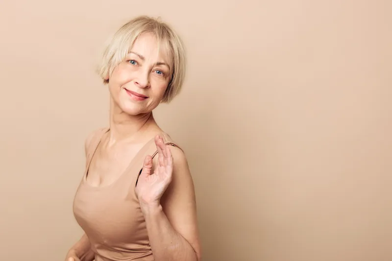 Smiling mature woman with short blonde hair wearing sleeveless beige top poses against matching background. Concept of confidence, aging gracefully, lifestyle, and natural beauty in studio.