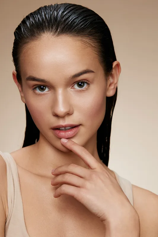 Young female model with wet hair and natural makeup, showcasing a fresh, glowing look against a soft beige background, ideal for beauty and skincare themes