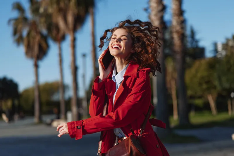 A young woman with curly hair in a red jacket chats on her phone, smiling while strolling along a palm lined street under clear blue skies.