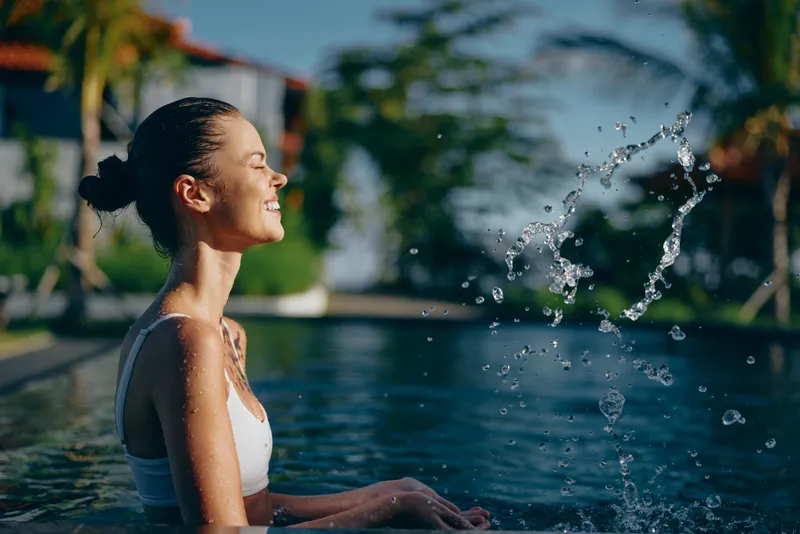 Joyful woman enjoys a refreshing moment by a sunny pool, smiling as water arcs from the edge, embodying casual lifestyle, outdoor relaxation and summer leisure in a serene poolside scene
