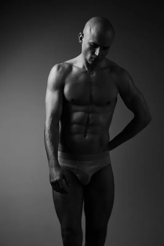 Black and white image of a muscular man posing gracefully, showcasing strength, confidence, and vulnerability in a simple yet captivating composition.