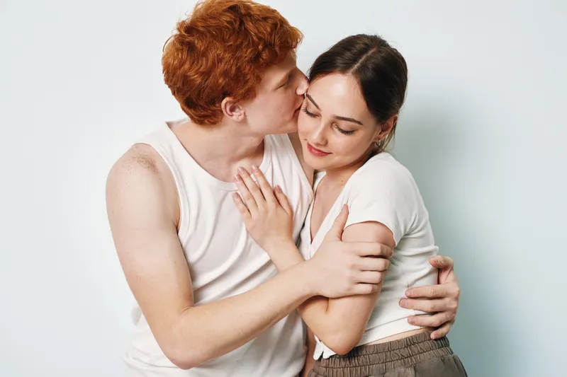 Young couple embracing indoors with affection, man kissing woman on cheek, casual clothing and warm emotional connection, love and tender moment captured in studio.