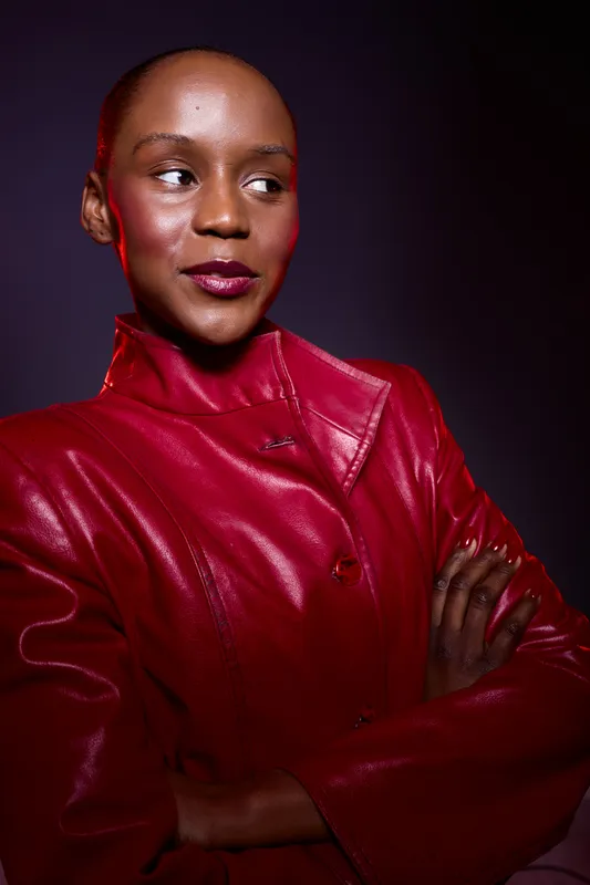 Confident dark skinned African woman wearing a red leather jacket stands with crossed arms against dark background. Studio portrait with dramatic lighting and strong expression.