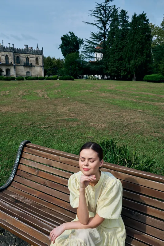 Woman sits on a bench in a park, outdoor scene with nature and castle in distance, serene and peaceful contemplation in a calm landscape with soft light and quiet atmosphere