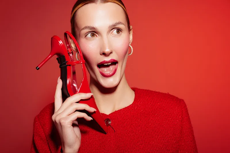 Young woman in red sweater holds red high heel shoe near face with surprised expression on red background. Concept of fashion, style, footwear, and vibrant color contrast.