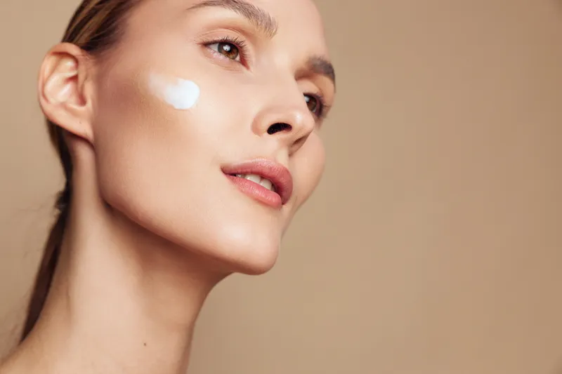 Closeup of young woman with smooth skin applying moisturizer cream on cheek, natural makeup, soft lighting, neutral background, skincare beauty routine concept.